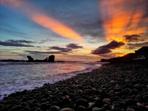 El Salvador: Cultural Immersion Tour With Workshops And More A scenic shot of a beach with smooth rocks in the foreground, waves crashing on the shore, and a stunning sunset casting orange rays through the clouds.