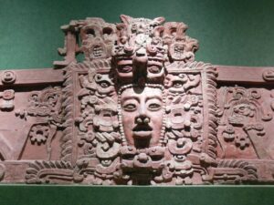 El Salvador In And Out: San Salvador, El Boqueron And Mayan History At Tazumal (4 days) This image shows a detailed reddish-brown stone carving featuring Mayan motifs, including faces and intricate symbolic patterns, set against a green background.