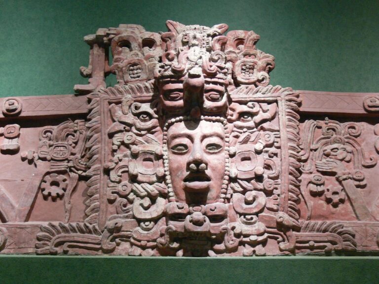 This image shows a detailed reddish-brown stone carving featuring Mayan motifs, including faces and intricate symbolic patterns, set against a green background.
