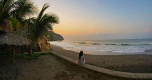 El Salvador Short And Sweet (Six Days) – Beach Edition A woman sits on a wall overlooking a dark sand beach at sunset, framed by palm trees and a thatched-roof structure.