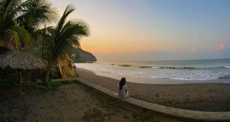 A woman sits on a wall overlooking a dark sand beach at sunset, framed by palm trees and a thatched-roof structure.