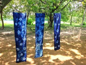 Suchitoto Indigo Dyeing Workshop Three blue, tie-dyed fabrics hang on a clothesline between trees in a sun-dappled yard, showcasing the unique circular and spiral patterns of the dye.