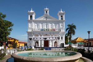 Essential El Salvador: Nine Days In The Land Of Volcanoes A white colonial church with towers and "Suchitoto" sign is seen behind a fountain in a town square setting on a sunny day.