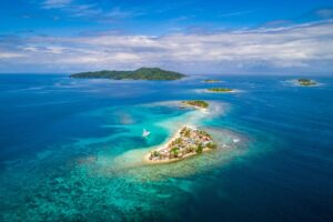 Discover The Beauty Of El Salvador And Honduras Aerial view of a tropical archipelago with clear turquoise waters, featuring a small island densely populated with huts and a boat anchored nearby.