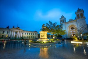 A Quick Taste Of El Salvador: San Salvador, Coffee And Ruta De Las Flores (4 days) A captivating nighttime shot of a city square featuring a statue, historic buildings, and a cathedral reflecting in the wet pavement, illuminated by streetlights.
