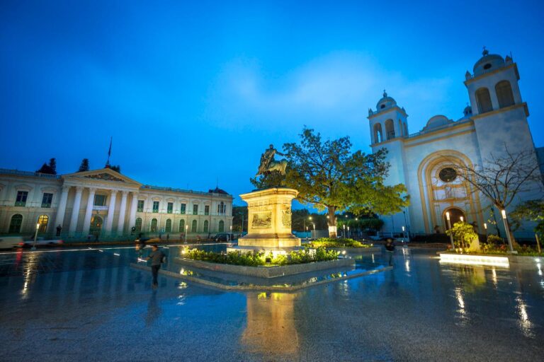 A captivating nighttime shot of a city square featuring a statue, historic buildings, and a cathedral reflecting in the wet pavement, illuminated by streetlights.