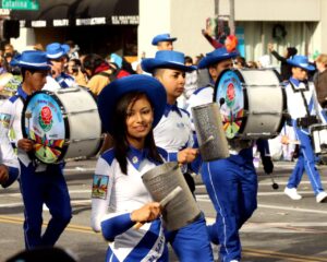 San Salvador City Tour And El Boqueron Visit A marching band dressed in blue and white uniforms parades down a street, with some members playing drums and others shaking metal percussion instruments.
