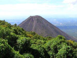 El Salvador Short And Sweet (Six Days) A volcano rises majestically in the center, flanked by lush green vegetation, with a distant view of landscape under a clear sky.