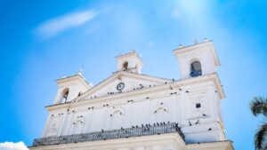 Cultural And Natural Highlights Of El Salvador (11 days) An upward shot of a white building with a clock and towers against a bright blue sky.