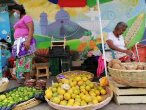 One Week In El Salvador: Option 1 A vibrant market scene unfolds with two women amidst baskets of citrus fruits and potatoes, against a backdrop of a colorful mural.