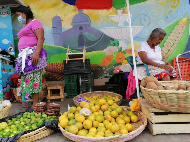 A vibrant market scene unfolds with two women amidst baskets of citrus fruits and potatoes, against a backdrop of a colorful mural.