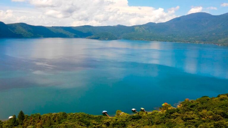 A scenic view of a turquoise lake surrounded by lush green mountains under a partly cloudy sky.