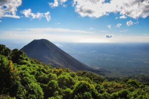 Volcano Tour In El Salvador (One Week) A volcano rises majestically amidst lush greenery under a bright blue sky with scattered clouds.