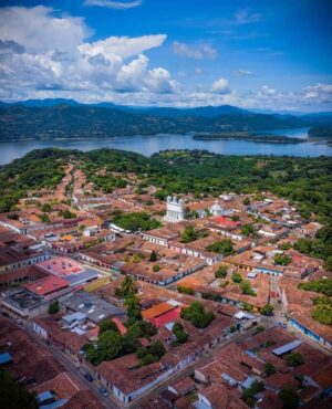 Extended Tour In El Salvador, Central America’s Little Pearl (18 days) An aerial view captures a town with red-tiled roofs nestled among lush green trees, with a lake and mountains in the distance under a cloudy blue sky.