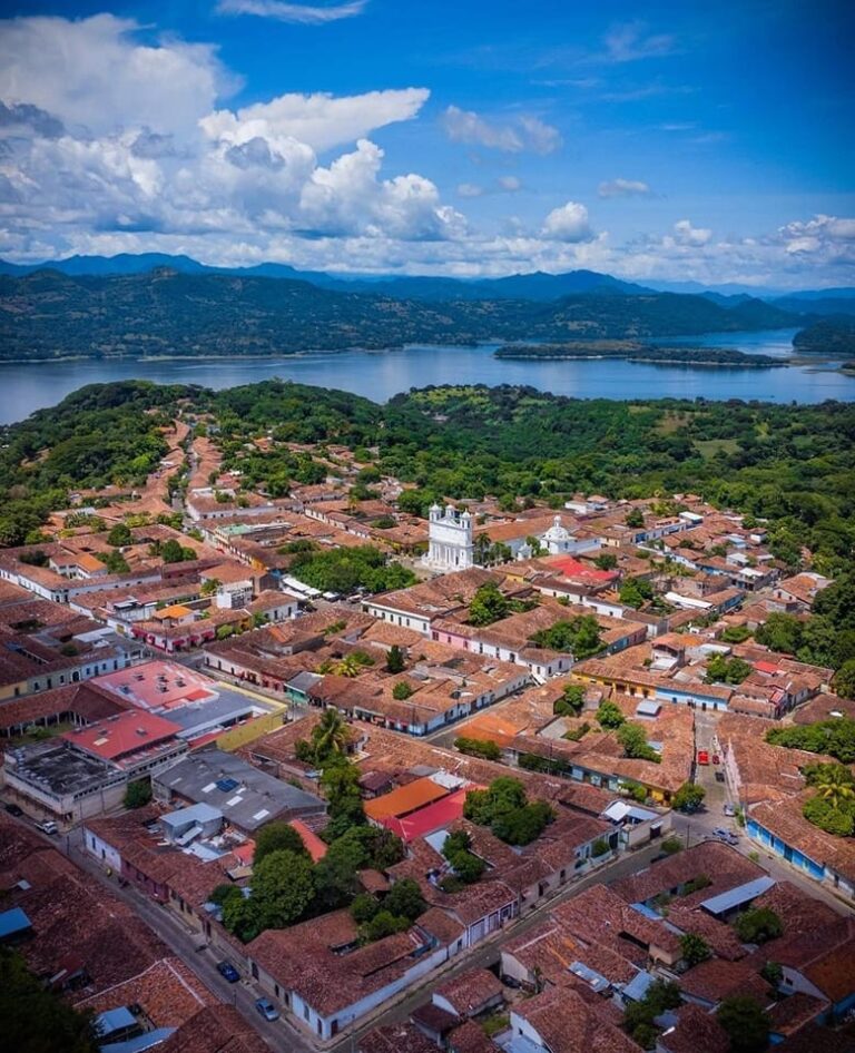 An aerial view captures a town with red-tiled roofs nestled among lush green trees, with a lake and mountains in the distance under a cloudy blue sky.
