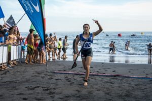 Surf City El Salvador A female athlete in a USA uniform runs barefoot on a beach with a paddle in hand, triumphantly raising a finger, while a crowd cheers behind barriers and other competitors paddle in the sea.