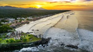 El Salvador’s Magnificent Beaches An aerial shot of a tropical beach at sunset with mountains in the distance and a village next to the coastline.