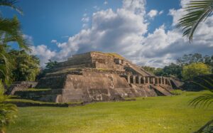 Eleven Best Travel Activities In El Salvador A stepped pyramid rises from a grassy field under a cloudy sky, framed by lush greenery on either side.