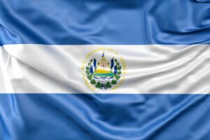 Traveling To El Salvador: Key Information This image shows the flag of El Salvador, which consists of two horizontal blue stripes and a central white stripe, with the national coat of arms centered in the white stripe.