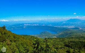 What To Visit During Your Travel To El Salvador? A breathtaking aerial view of a serene blue lake surrounded by lush green mountains under a clear blue sky.