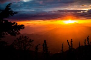 Welcome To El Salvador: Country Of Bitcoin, Surfing And Volcanoes! A vibrant sunset over a mountainous landscape, with silhouettes of trees and foliage in the foreground, and power lines stretching across the fiery sky.