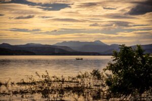 Lakes Of El Salvador A serene landscape featuring a tranquil lake under a cloudy, golden sky, with distant mountains and a small boat, framed by lush vegetation in the foreground.